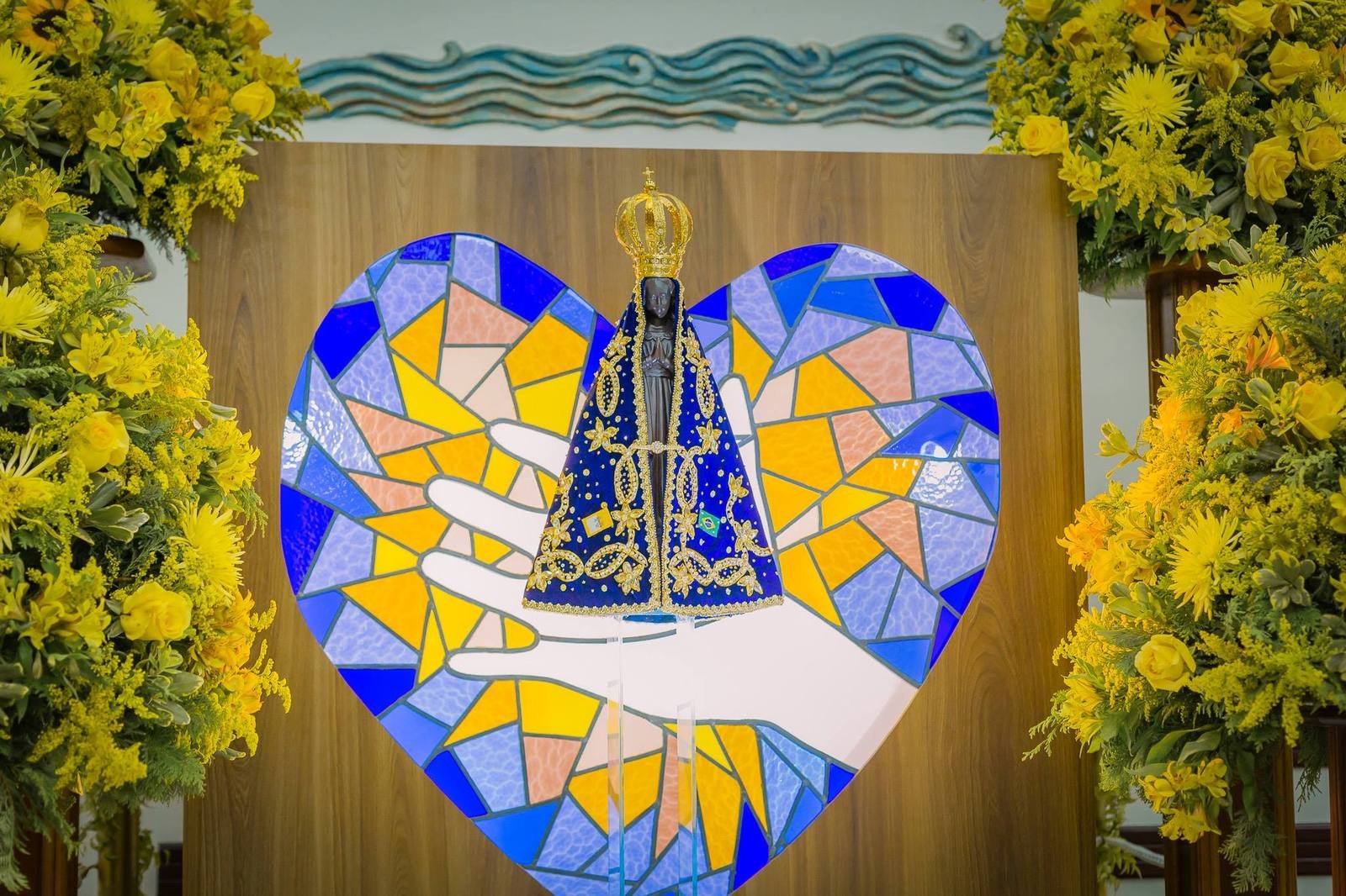 Imagem de Nossa Senhora Aparecida sobre um coração em vitral azul e amarelo, entre flores amarelas